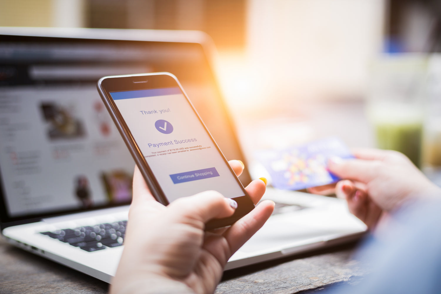 Person holding a smartphone with a payment confirmation screen, with a laptop and credit card in the background.