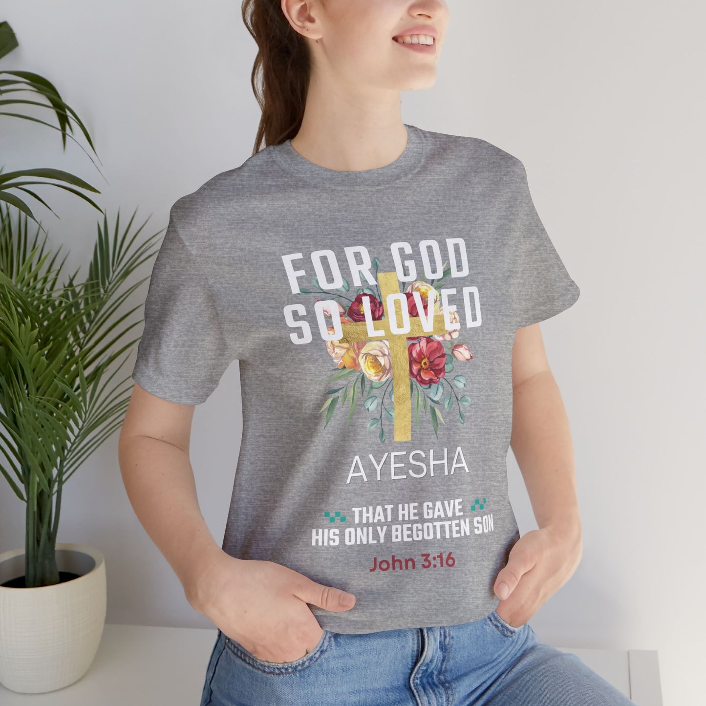 A woman wearing a heather gray graphic t-shirt with a floral cross design and the text 'FOR GOD SO LOVED AYESHA THAT HE GAVE HIS ONLY BEGOTTEN SON John 3:16'.