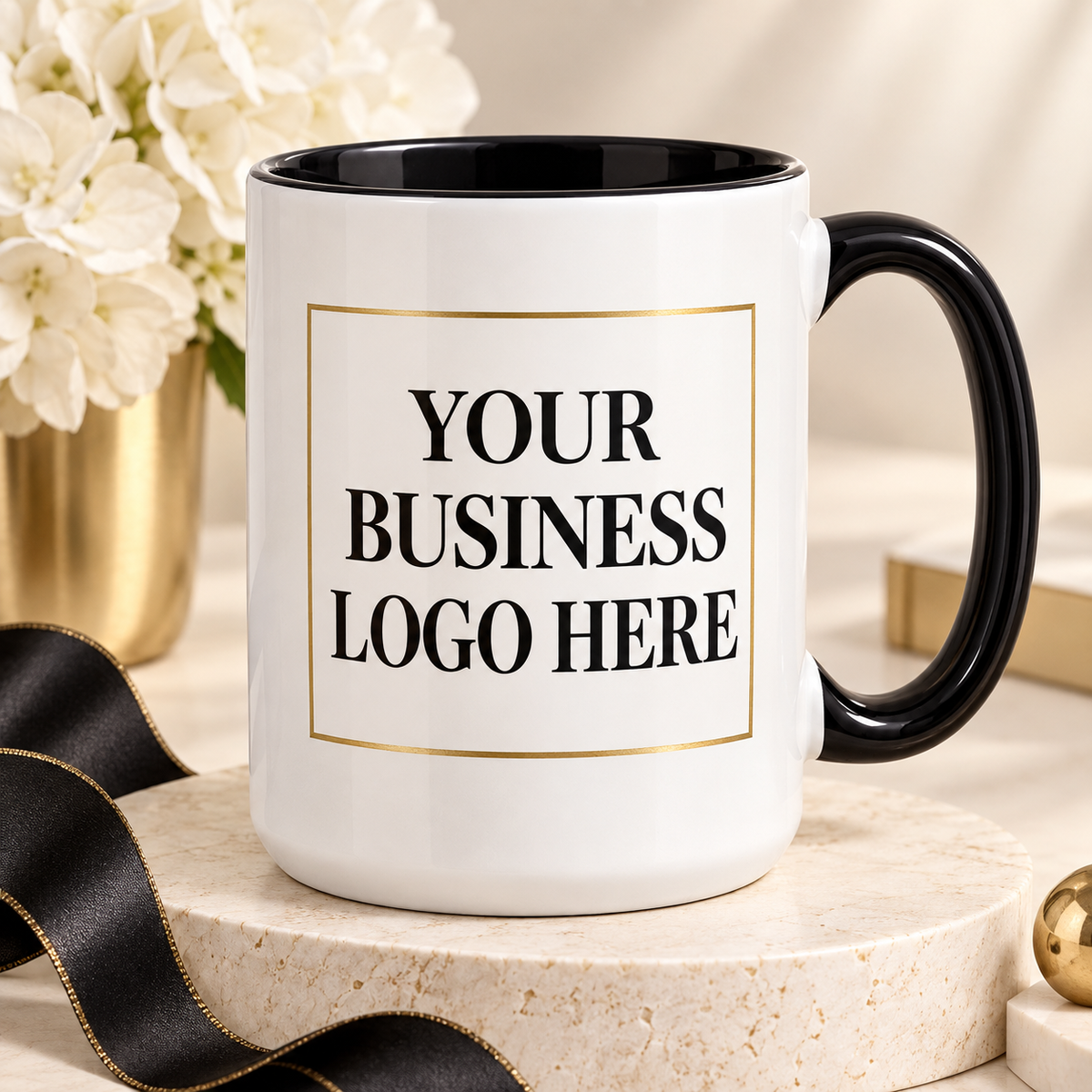 A white ceramic mug with a black handle and black interior, featuring a customizable gold-bordered placeholder box with the text 'YOUR BUSINESS LOGO HERE'. The mug is styled on a round stone pedestal with a black ribbon, white flowers, and a neutral background.